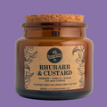 An amber glass jar candle with a cork stopper and a kraft paper label reading 'Rhubarb & Custard' by Little Earth Co, photographed on a light gray background.