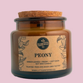 An amber glass jar candle with a cork stopper and a kraft paper label reading 'Little Inferno' and 'Peony' on a light gray background.