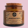 An amber glass jar candle with a cork stopper and a tan paper label reading HONEYSUCKLE by Little Meowz, photographed against a plain light gray background.