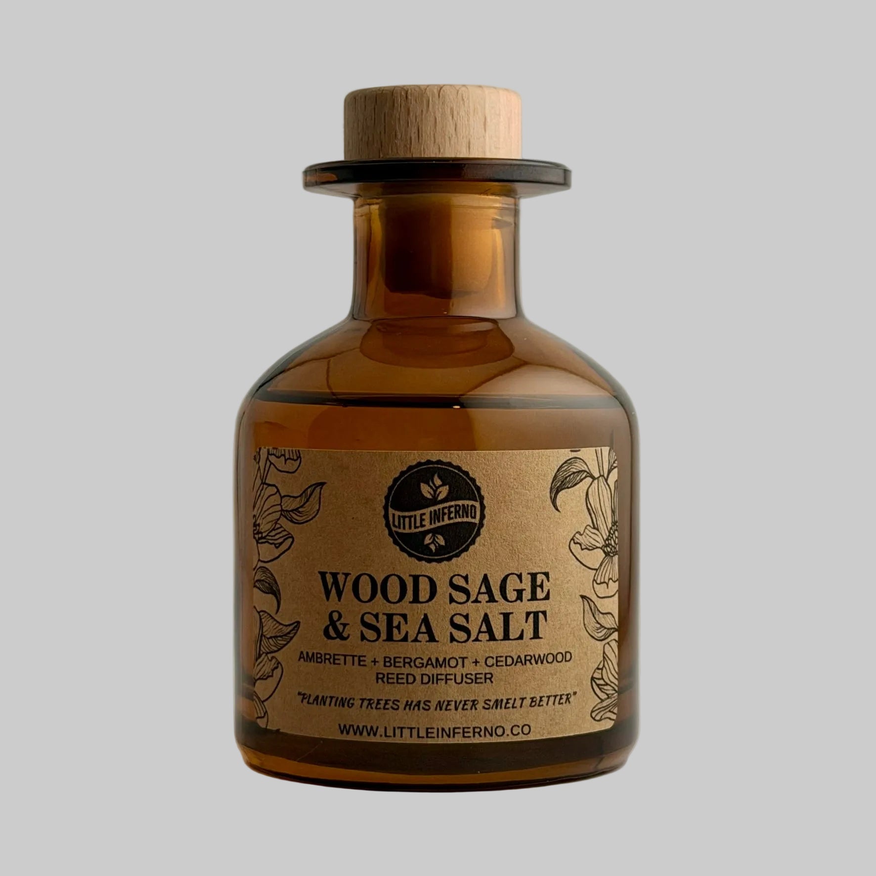 An amber glass bottle with a natural wood cork stopper and a kraft paper label reading 'Wood Sage & Sea Salt' by Little Inferno, photographed against a plain light gray background.