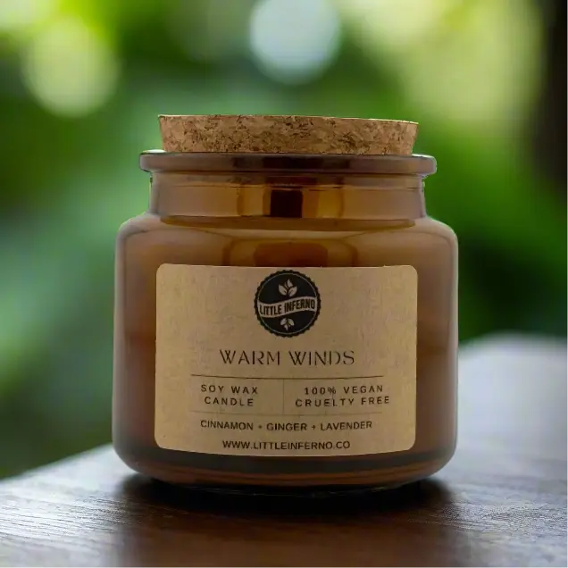 Candle jar with cork lid and 'Little Inferno' label on a wooden surface with a blurred green background