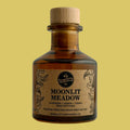 A small amber glass bottle with a light wood cork stopper and a kraft paper label reading 'MOONLIT MEADOW' by Little Emperor, photographed against a plain light gray background.