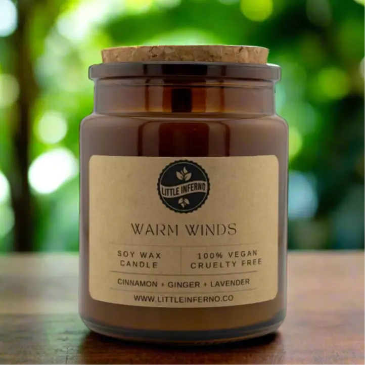 Scented candle labeled 'Warm Winds' by Little Inferno on a wooden surface with a blurred green background.